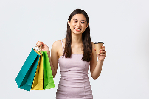 Lifestyle, shopping and tourism concept. Carefree happy asian woman in dress, drinking coffee on her way to next shop, holding bags with new clothes, enjoying weekend in mall, white background.
