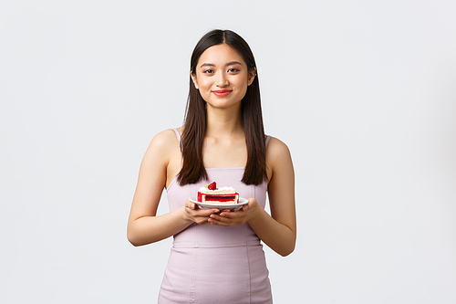Lifestyle, holidays, celebration and food concept. Beautiful asian girl in evening dress enjoying eating sweet delicious piece cake, smiling delighted, standing white background.