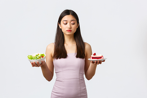 Healthy lifestyle, leisure and food concept. Beautiful asian girl trying resist temptation to bite sweet delicious cake full of calories, staying on diet and eating salad, white background.