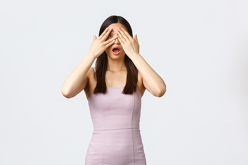 Luxury women, party and holidays concept. Excited attractive asian birthday girl in elegant dress, close eyes with palms and peek through fingers, awaiting for surprise gift, white background.