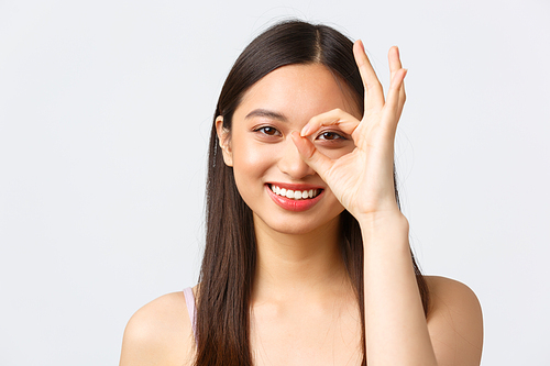 Beauty, fashion and people emotions concept. Close-up portrait of kawaii happy asian girl, smiling white teeth and showing okay gesture over eye, standing white background cheerful.