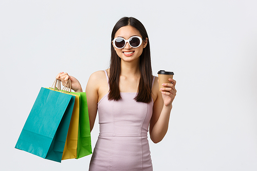 Lifestyle, leisure and tourism concept. Elegant and stylish asian woman in sunglasses and dress, drinking coffee and holding shopping bags, buying new clothes for summer vacation.