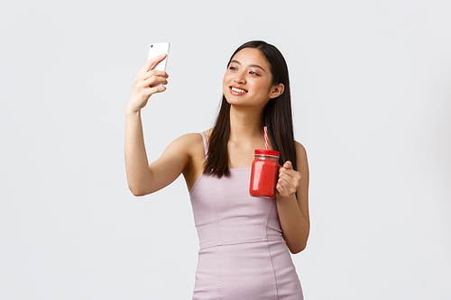 Healthy lifestyle, people emotions and leisure concept. Attractive asian girl blogger in dress, taking selfie with smoothie and smiling at mobile phone camera, white background.