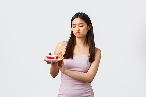 Lifestyle, holidays, celebration and food concept. Reluctant and upset, gloomy asian woman in dress, refusing eat cake, showing stop gesture and looking at plate with regret, white background.