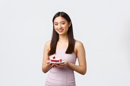 Lifestyle, holidays, celebration and food concept. Beautiful asian woman in party dress, enjoying celebrating friend birthday, get piece cake and smiling camera, standing white background.