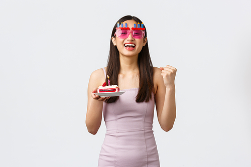 Celebration, party and holidays concept. Optimistic happy beautiful asian girl in funny glasses and dress, fist pump in triumph, encourage herself on positive energy, making wish at birthday cake.