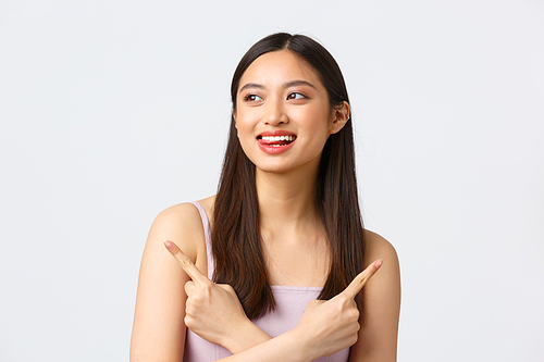 Beauty, fashion and people emotions concept. Dreamy happy beautiful asian woman in dress, smiling and lick lips from temptation, pointing fingers sideways two choices, white background.