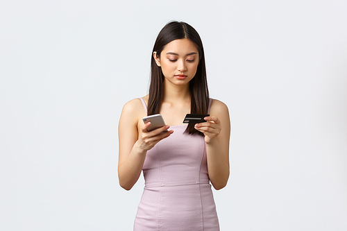 Luxury women, party and holidays concept. Serious-looking beautiful asian girl in evening dress making online order, holding credit card and smartphone, paying for delivery, white background.