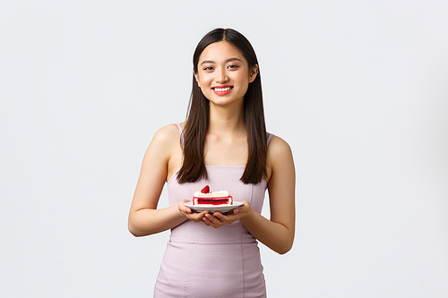 Lifestyle, holidays, celebration and food concept. Beautiful smiling asian woman in party dress, holding plate with sweet cake and gazing camera, standing white background.