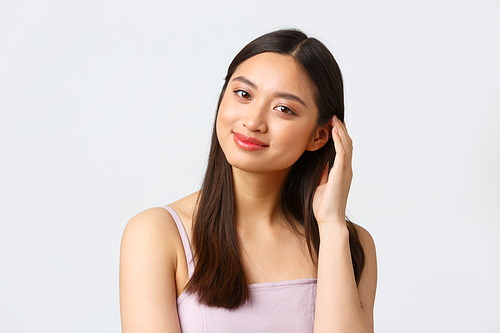 Beauty, fashion and people emotions concept. Close-up portrait of adorable young asian woman with perfect skin, smiling camera and brushing hair, tilt head coquettish, white background.