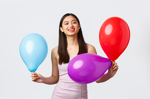 Celebration, party and holidays concept. Upbeat happy smiling asian woman in evening dress, laughing cheerful, celebrating birthday, holding colored balloons, white background.