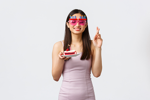 Celebration, party and holidays concept. Dreamy hopeful asian birthday girl in dress, cross fingers wish closed eyes and smile, holding piece cake with it candle, standing white background.