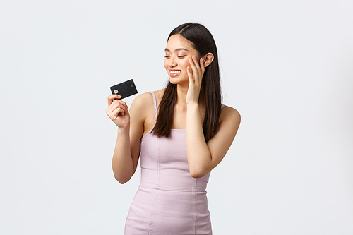 Luxury women, party and holidays concept. Pleased silly asian girl wasting all money, looking happy and satisfied at credit card after going shopping, wearing new evening dress, white background.