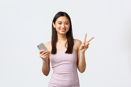 Luxury women, party and holidays concept. Lovely and cute asian girl in evening dress, showing peace sign and using mobile phone, smiling at camera, holding smartphone, white background.