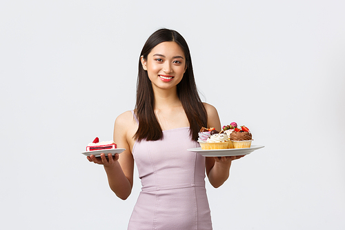 Lifestyle, holidays, celebration and food concept. Beautiful asian woman in dress holding plates with desserts, suggesting cake or plate with cupcakes, standing white background.
