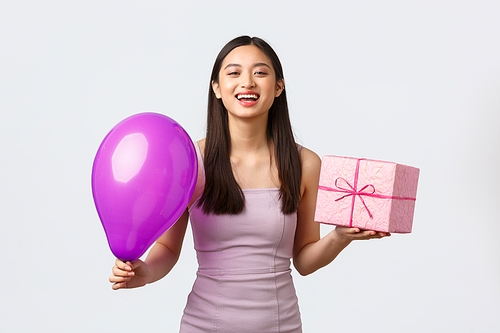 Celebration, party and holidays concept. Cheerful asian girl in evening dress, enjoying birthday, having fun, holding balloon and gift, laughing carefree, standing white background.