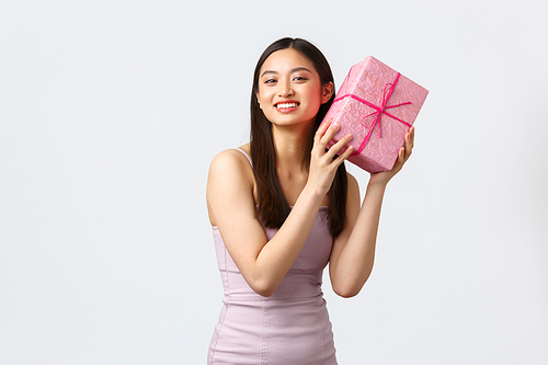 Celebration, party and holidays concept. Beautiful asian girl in evening dress, celebrating birthday, shaking b-day gift near ear to guess what inside, smiling happy, white background.