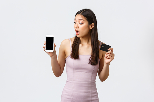 Luxury women, party and holidays concept. Surprised pretty asian woman in evening dress, looking curious at smartphone screen, showing credit card and mobile phone display, white background.