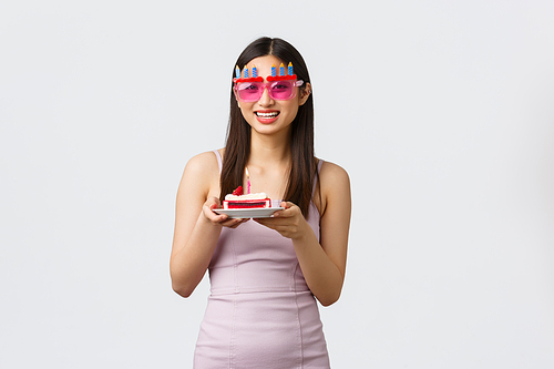 Celebration, party and holidays concept. Happy excited birthday girl in dress and funny sunglasses, holding piece cake with candle, making wish and having fun, standing white background.