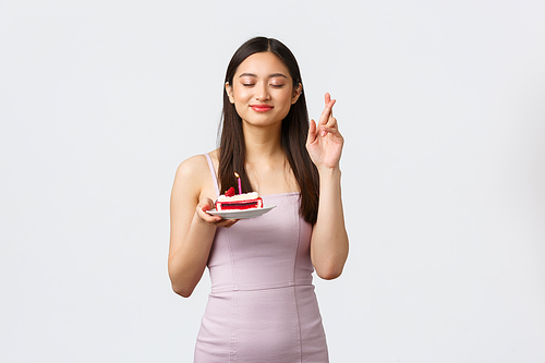 Celebration, party and holidays concept. Happy dreamy beautiful asian woman in dress, cross fingers good luck with closed eyes, smiling as making wish over birthday cake, white background.