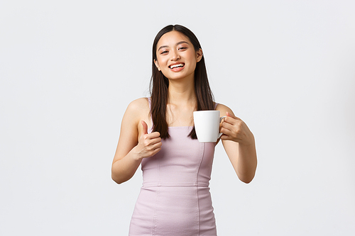 Lifestyle, people emotions and glamour concept. Gorgeous asian woman drinking coffee and showing thumbs-up, smiling pleased, enjoying drink as standing over white background.