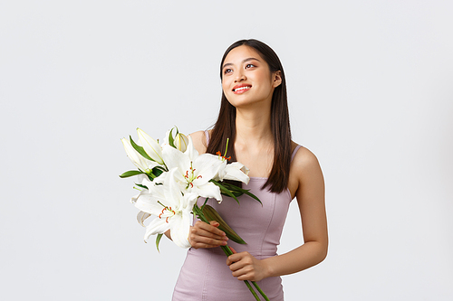 Holidays and events, celebration concept. Dreamy and romantic, happy smiling asian woman in stylish dress, looking upper left corner and holding bouquet of lilies, receive flowers as gift.