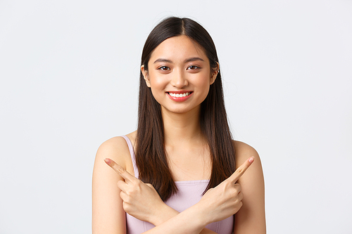 Beauty, fashion and people emotions concept. Close-up portrait of pretty asian girl in dress, pointing fingers sideways and smiling, suggesting both variants, two choices, standing white background.