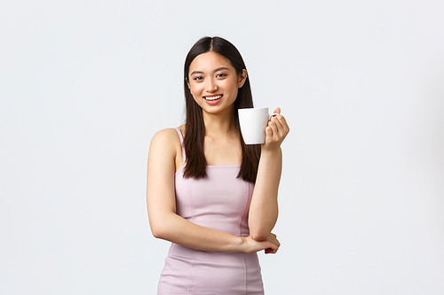Lifestyle, people emotions and glamour concept. Stylish good-looking young asian woman in trendy dress, smiling and having small-talk during lunch, drinking coffee in office, white background.