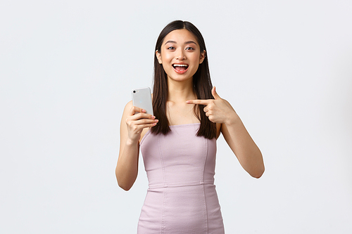 Luxury women, party and holidays concept. Excited happy asian girl in evening dress, pointing at mobile phone and looking enthusiastic, telling about awesome smartphone application.