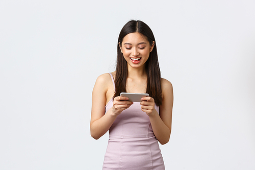 Luxury women, party and holidays concept. Happy carefree asian girl in evening dress, playing mobile game, holding smartphone horizontally and smiling at display, white background.