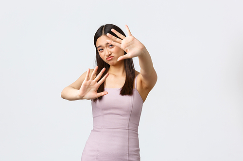 Luxury women, party and holidays concept. Stop photographing me. Displeased asian girl in evening dress extend hands to defend eyes from light of camera flesh, standing white background.