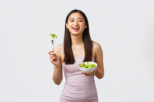 Healthy lifestyle, leisure and people emotions concept. Portrait of happy asian girl eating salad in beautiful evening dress, have bite of fresh vegetables, standing white background.