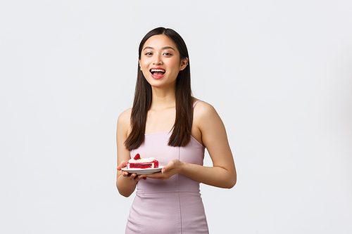 Lifestyle, holidays, celebration and food concept. Enthusiastic beautiful asian girl eating at party, wearing dress, holding plate with piece cake, laughing and smiling, white background.