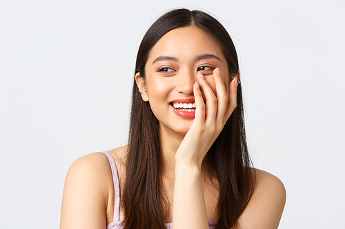 Concept of beauty, fashion and makeup products advertisement. Close-up portrait of adorable, romantic blushing asian girl, smiling and laughing coquettish, standing white background, looking left.