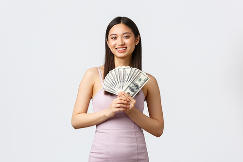 Luxury women, party and holidays concept. Beautiful smiling asian woman in evening dress, showing money, like shopping, standing white background delighted. Copy space