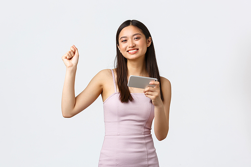 Luxury women, party and holidays concept. Cheerful pleased good-looking asian girl dancing like champion, winning in game, playing on mobile phone, feeling happy, white background.