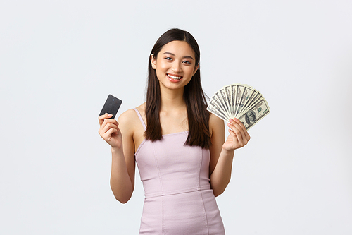 Luxury women, party and holidays concept. Gorgeous young successful asian woman in evening dress smiling, showing cash and credit card, suggest two ways of investments, white background.