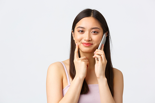 Beauty, fashion and people emotions concept. Close-up portrait of beautiful asian girl looking intrigued during phone call, talking on mobile and smiling intrigued, white background.