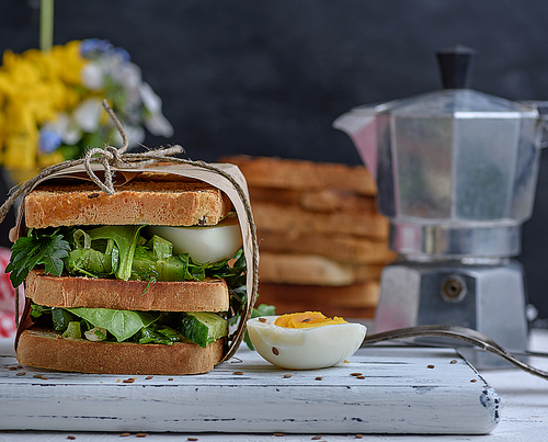 sandwich of French toast and lettuce leaves and boiled egg, a vegetarian food wrapped in paper on a white wooden board