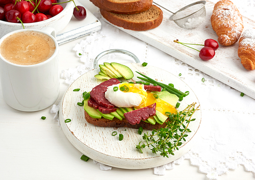 toast with poached egg and avocado on a round board, next to croissants and ripe red cherries, morning breakfast, top view on a white table