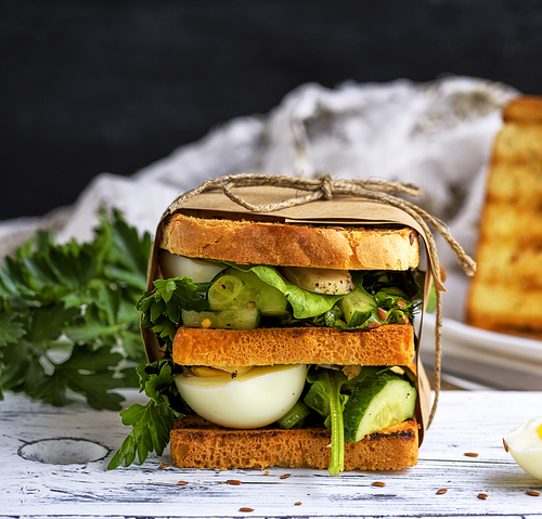 sandwich of French toast and lettuce leaves and boiled egg, a vegetarian food wrapped in paper on a white wooden board