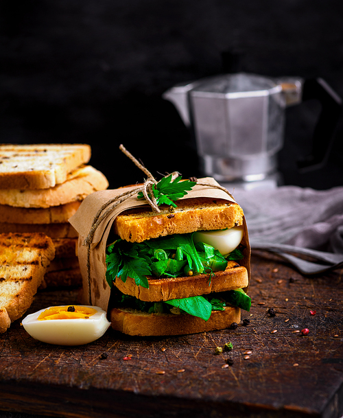 sandwich of French toast and lettuce leaves and boiled egg, a vegetarian food  wrapped in paper on a brown wooden board