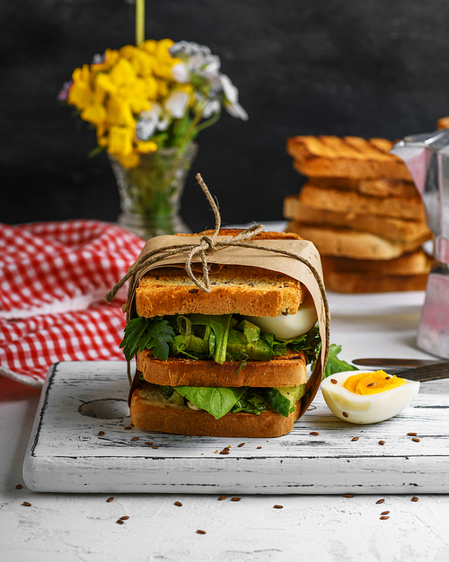 sandwich of French toast and lettuce leaves and boiled egg, a vegetarian food  wrapped in paper on a white wooden board