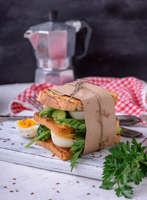 sandwich of French toast and lettuce leaves and boiled egg, a vegetarian food wrapped in paper on a white wooden board