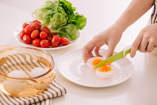 Close up of females hands cutting boiled egg for his breakfast and mixed vegetables on white wooden background