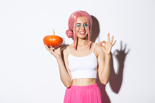 Image of happy girl celebrating halloween, wearing pink wig, holding pumpkin and showing okay sign, standing over white background.