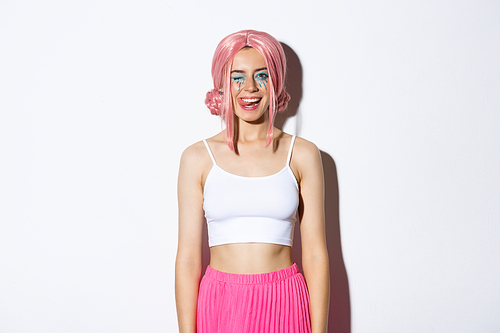 Portrait of carefree smiling female model in pink party wig, winking and showing tongue, celebrating halloween, standing over white background.
