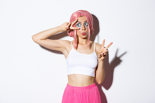 Image of silly beautiful female model with pink wig and halloween makeup, looking left surprised, making peace signs, standing over white background.