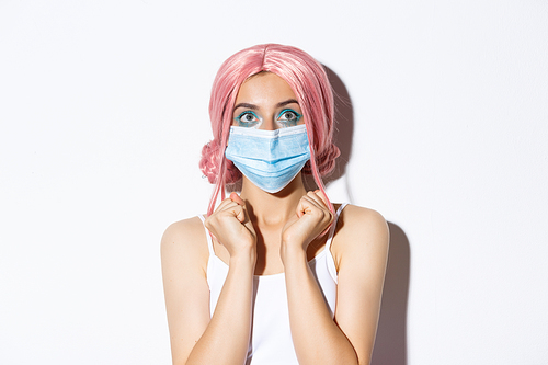 Covid-19 concept. Close-up of hopeful cute girl in medical mask, looking up excited, standing over white background in pink wig.