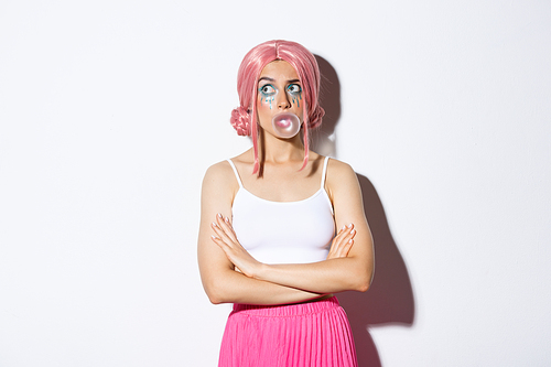 Image of confused party girl in pink wig, wigh bright makeup, blowing bubble gum and looking indecisive at upper left corner, standing over white background.
